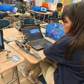 Student working on a robotics build next to a laptop, with tools and parts on a desk.