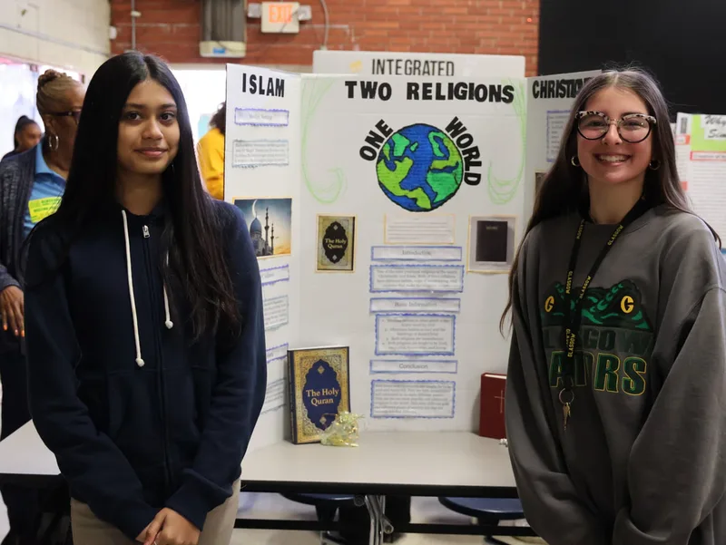 Two students standing by a display board about Islam and Christianity at a school project fair.

