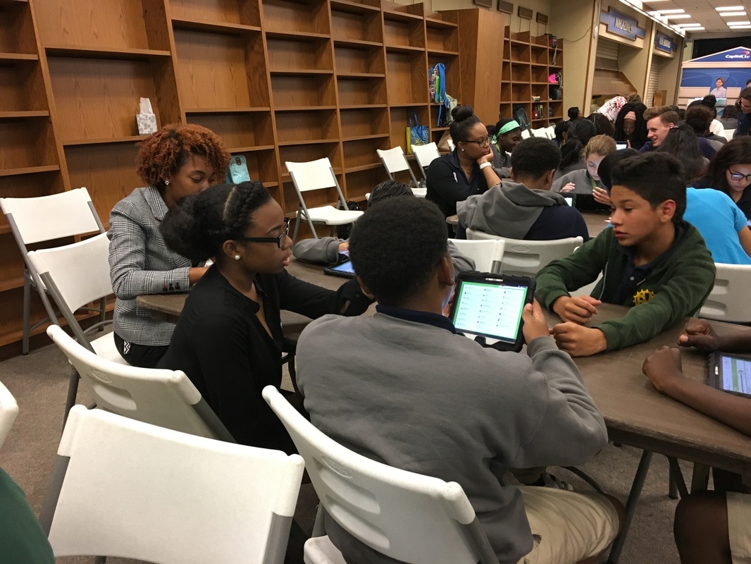 A group of students seated at a table with a volunteer. The students are working with tablets. 