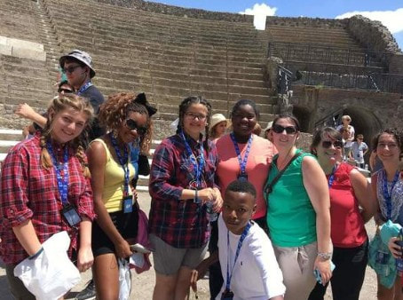 Group of students and adults pose in stone amphitheater with high steps in background.

