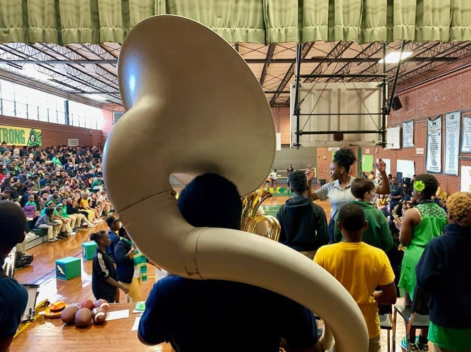 Student holding a tuba and standing before a large group of students in a gymnasium