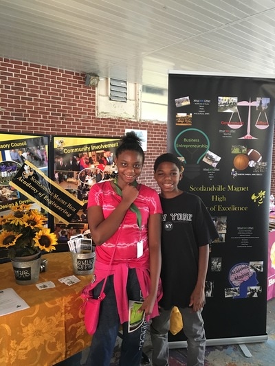 Two students smile at the camera. They are posing in front of a local high school's display. 