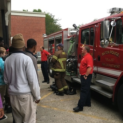 A group of students standing outside in front of a fire truck. Three firefighters are speaking to them. 