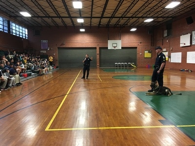 A group of students seated in the gym bleachers. Two police officers are speaking to them. One police officer has a dog with him. 