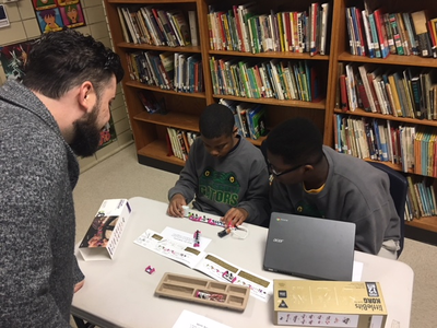 A volunteer assists two male students working with a laptop and robotics equipment.